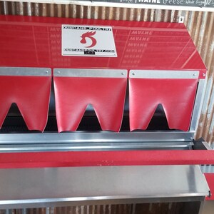 May include: Red metal chicken feeder with three red flaps and a silver tray. The feeder has a sign that says "Automatic Poultry Feeder".