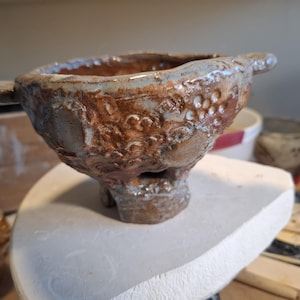 Può includere: Una ciotola in ceramica fatta a mano con una consistenza rustica e irregolare e una smaltatura lucida nei toni del marrone e del grigio. La ciotola presenta due piccole maniglie laterali, un'ampia apertura e una base spessa a piedistallo.