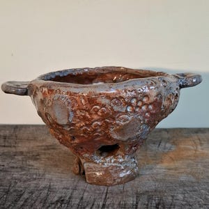 Op de afbeelding: Een kleine, bruine keramische kom met twee handvatten en een getextureerd oppervlak. De kom heeft een ruwe, ongelijke afwerking en lijkt handgemaakt te zijn.