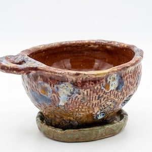 May include: A small, round, hand-formed ceramic bowl with a single side handle and a matching saucer base. The exterior features a textured, cross-hatched pattern in shades of brown, blue, and white, while the interior has a smooth, glossy brown finish.