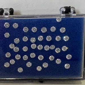 May include: A rectangular clear plastic display case containing numerous small, round, faceted, colorless stones arranged on a vibrant blue foam insert. The stones exhibit a brilliant, reflective quality, and the case features black hinges at the top.