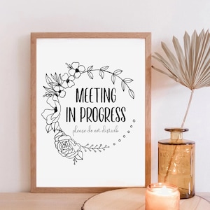 May include: A framed print with the text "MEETING IN PROGRESS please do not disturb" encircled by a floral wreath. The frame is light brown. A vase with a dried plant and a lit candle are in the background.