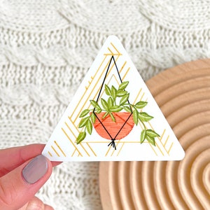 May include: A triangular sticker featuring a watercolor illustration of a hanging plant. The plant is in a terracotta-colored pot, suspended by black macrame cords, with green leaves. The background has a gold geometric pattern.