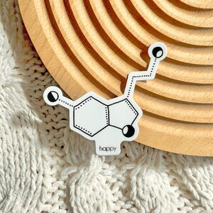 Puede incluir: Una pegatina blanca con un dibujo de líneas negras de una estructura molecular química. El diseño incluye una configuración central de anillos hexagonales y pentagonales con líneas ramificadas que terminan en pequeños círculos. La palabra happy está impresa en negro.