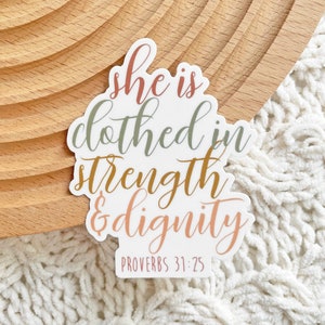 May include: White sticker with the text "She is clothed in strength & dignity" in a cursive font with different colored words. The text "Proverbs 31:25" is printed below the main text.