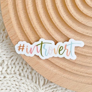 May include: A white sticker with the text "#introvert" written in colorful script. The sticker is on a light brown wooden surface.