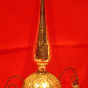 Peut inclure: Un sommet d'arbre de No&euml;l vintage en verre avec une finition argent&eacute;e et un motif en maille de fil. Le sommet a trois cloches attach&eacute;es &agrave; lui avec des cordons noirs.