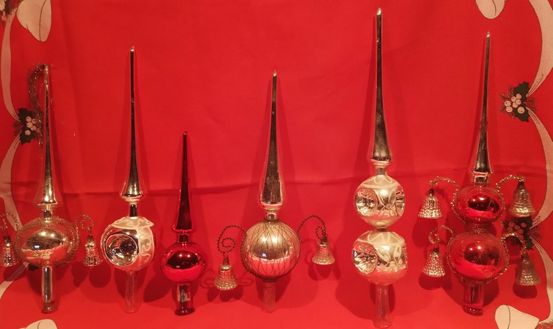 Peut inclure: Six d&eacute;corations de sommet d'arbre de No&euml;l en verre vintage dans diff&eacute;rents styles. Les sommets sont en verre argent&eacute;, rouge et transparent avec des cloches et un sommet pointu. Les sommets sont affich&eacute;s sur un fond rouge.