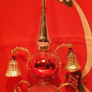 Peut inclure: Un sommet d'arbre de No&euml;l vintage en verre rouge avec une fl&egrave;che en m&eacute;tal argent&eacute; et quatre cloches. Le sommet est d&eacute;cor&eacute; de paillettes dor&eacute;es et a une base en verre rouge.