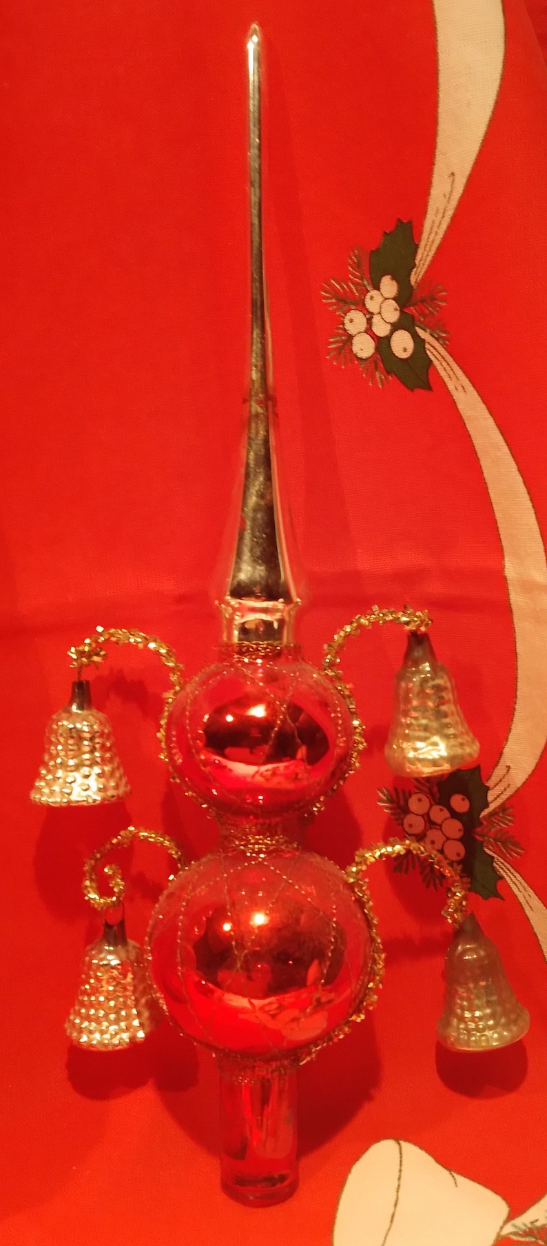Peut inclure: Un sommet d'arbre de No&euml;l vintage en verre rouge avec une fl&egrave;che en m&eacute;tal argent&eacute; et quatre cloches. Le sommet est d&eacute;cor&eacute; de paillettes dor&eacute;es et a une base en verre rouge.