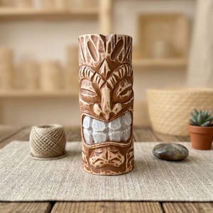 May include: A brown, carved tiki mug with a detailed face design. The mug features a wide, toothy grin and is set on a wooden table. A small succulent in a terracotta pot and a stone are also visible.