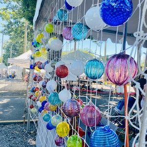 Op de afbeelding: Een display van kleurrijke, ronde glazen ornamenten die aan een wit net hangen. De ornamenten zijn in verschillende kleuren, waaronder blauw, groen, geel, wit en paars. Sommige hebben een textuur, andere zijn glad. De ornamenten zijn opgehangen met witte touwtjes.