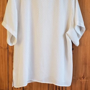 May include: White short-sleeved blouse with a small black polka dot pattern. The blouse has a round neckline and is hanging on a wooden hanger. The label reads "2H Collection".
