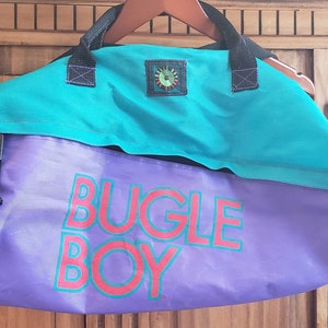 May include: A purple and teal duffel bag with the words "Bugle Boy" printed in red and white on the front.