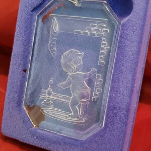 May include: A clear glass Christmas ornament featuring an etched design of a child standing in front of a fireplace. The ornament is in a purple velvet box with the text "Etchings by Comar" on the box.