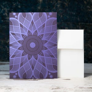 May include: A purple card with a detailed mandala design printed on it. The card is standing upright on a white wooden surface next to a white envelope.