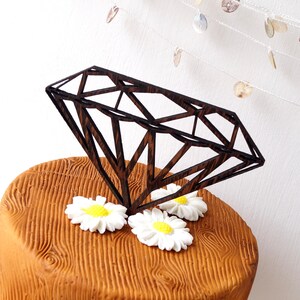 Pode incluir: Um bolo castanho com textura de madeira é decorado com margaritas brancas e um grande topo em forma de diamante preto.