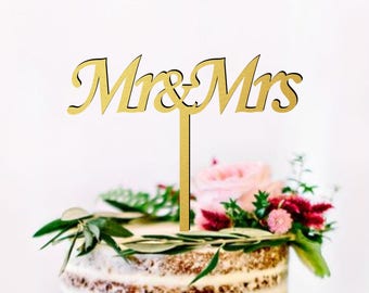 May include: A gold wooden cake topper with the text "Mr & Mrs" in a cursive font. The topper is placed on a two-tiered wedding cake decorated with greenery and flowers.