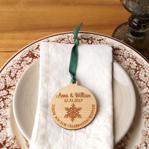 May include: A round wooden ornament with a snowflake design and the text "Anna & William 12.31.2017 Thank you for celebrating with us." The ornament is on a white napkin and a brown and white patterned plate.