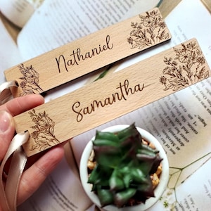 May include: Two wooden bookmarks with floral designs and personalized names, "Nathaniel" and "Samantha", are shown against a book background. The bookmarks have a light brown color and a satin ribbon.