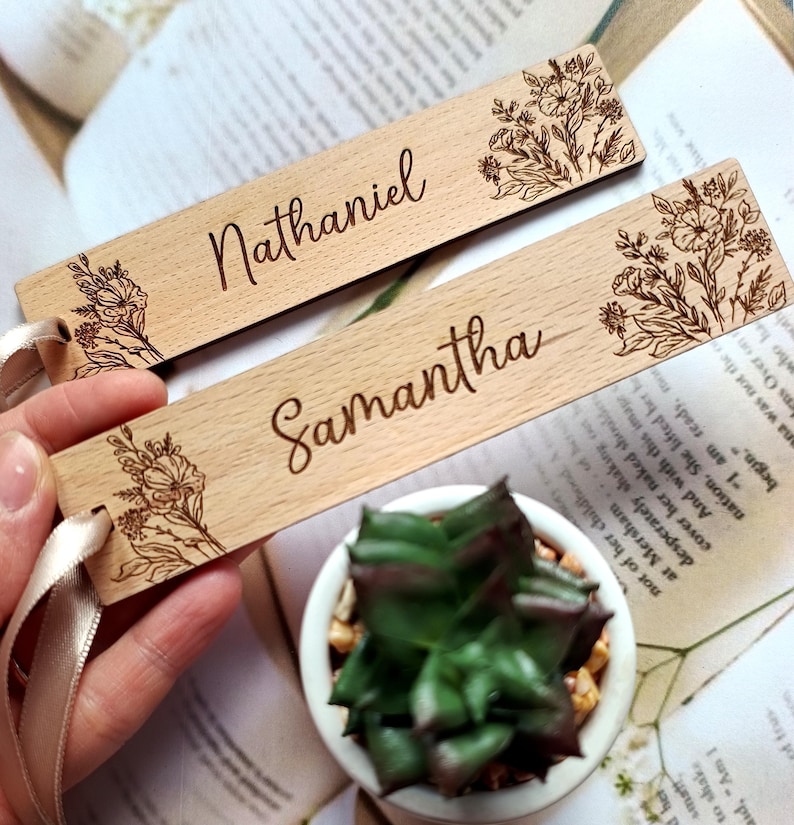 May include: Two wooden bookmarks with floral designs and personalized names, "Nathaniel" and "Samantha", are shown against a book background. The bookmarks have a light brown color and a satin ribbon.