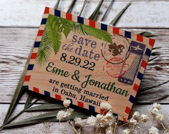 May include: A wooden save the date card with a tropical theme. The card is decorated with green leaves and a red, white, and blue striped border. The text on the card reads "Save the date 8.29.22 Esme & Jonathan are getting married in Oahu, Hawaii".