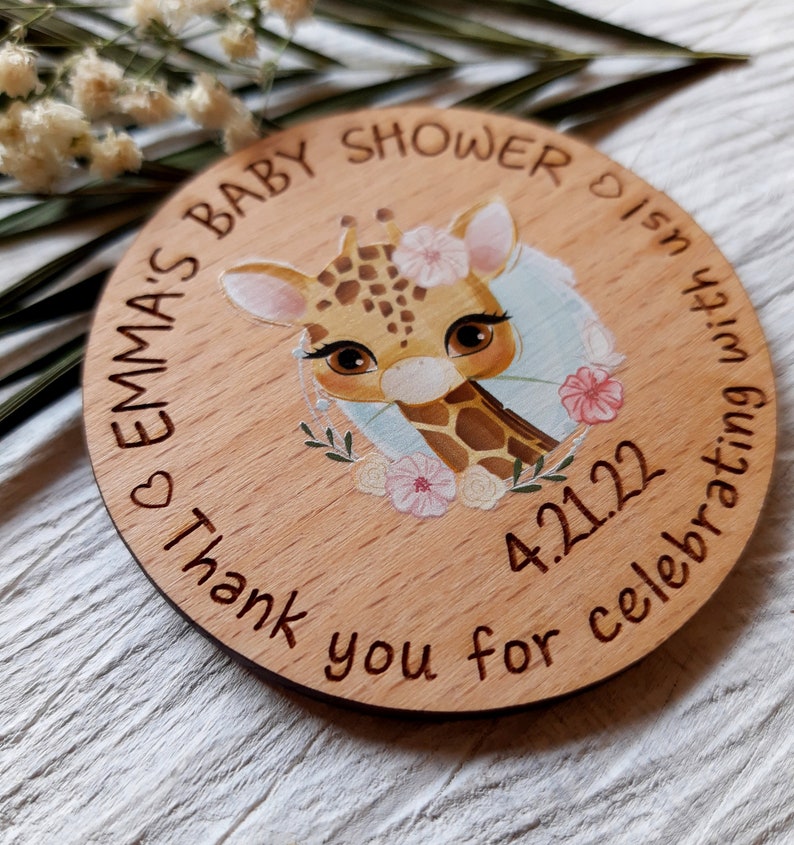 Peut inclure: Une pancarte ronde en bois avec une illustration de girafe, des fleurs et le texte "Emma's Baby Shower is in celebrating with 4.2.122 Thank you for celebrating with us".