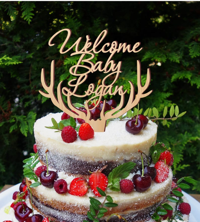 May include: A wooden cake topper with antlers and the text "Welcome Baby Logan". The topper is on a two-tiered cake decorated with cherries, raspberries, and strawberries.