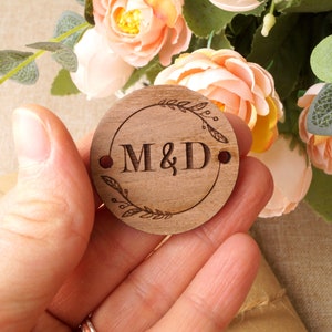 Op de afbeelding: Een ronde houten schijf met een bloemmotief en de initialen "M&D" gegraveerd in het midden. De schijf wordt in een hand gehouden.