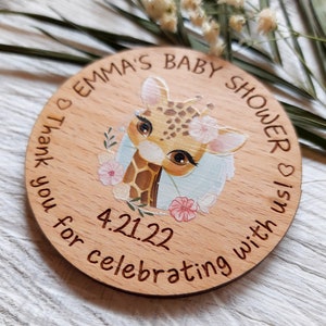 Peut inclure: Une faveur de douche de b&eacute;b&eacute; ronde en bois avec une illustration de girafe et des fleurs roses. Le texte indique "Emma's Baby Shower is on 4.21.22 Thank you for celebrating with us".
