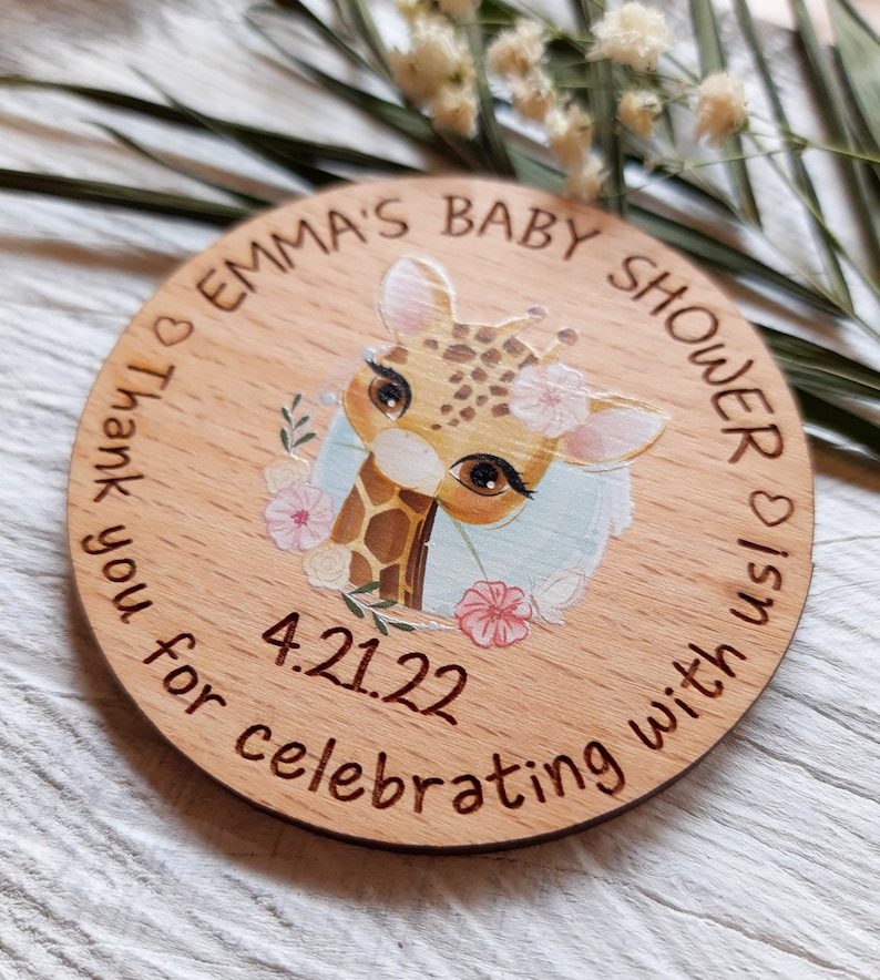 Peut inclure: Une faveur de douche de b&eacute;b&eacute; ronde en bois avec une illustration de girafe et des fleurs roses. Le texte indique "Emma's Baby Shower is on 4.21.22 Thank you for celebrating with us".