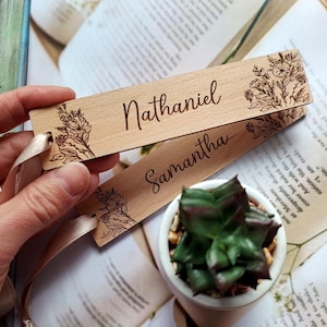 May include: Two wooden bookmarks with floral designs and personalized names, "Nathaniel" and "Samantha", are shown on a book with a small potted succulent plant.