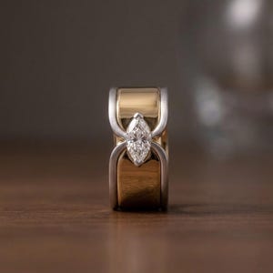 Könnte beinhalten: Ein breiter Ring mit einem polierten Mittelteil aus Gelbgold, der von zwei geschwungenen, silberfarbenen Metallbändern flankiert wird. Ein einzelner, klarer Edelstein im Marquise-Schliff ist vertikal in der Mitte mit silberfarbenen Krappen gefasst.