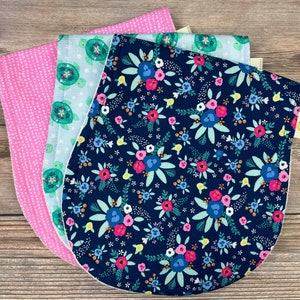 May include: Three baby bibs with different patterns are stacked on a wooden surface. The bibs feature a pink polka dot pattern, a light blue pattern with turtles, and a navy blue floral pattern with pink and blue flowers.