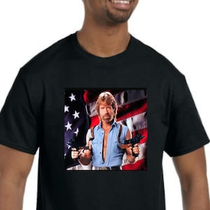 May include: Black t-shirt with a graphic of a man holding two guns in front of an American flag.