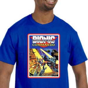 May include: Royal blue t-shirt featuring a graphic from "Bionic Commando." The graphic has a red border and displays the title in white block letters. The image shows a futuristic battle scene with soldiers and robots.