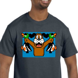 May include: A dark blue t-shirt featuring a pixelated image of a brown dog holding two green ducks in its mouth. The dog is wearing a red collar and has a white nose. The image is set against a blue background.