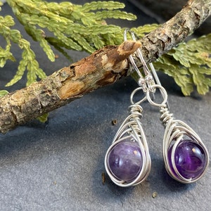 May include: A pair of silver wire wrapped earrings with amethyst gemstones. The earrings are dangling and have a teardrop shape.