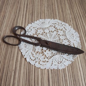 May include: A pair of vintage, dark brown metal scissors with two oval finger loops and a central pivot screw. The scissors are positioned on top of a round, white, intricate lace doily featuring a detailed floral and leaf pattern design.