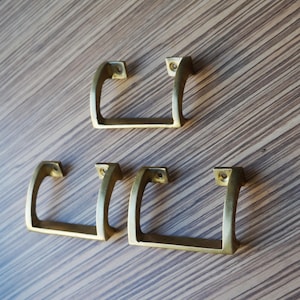 Peut inclure: Quatre poignées de meubles de couleur laiton avec un design carré et incurvé. Les poignées sont disposées sur une surface claire à grain de bois brun. Chaque poignée a une base rectangulaire avec deux trous de vis.