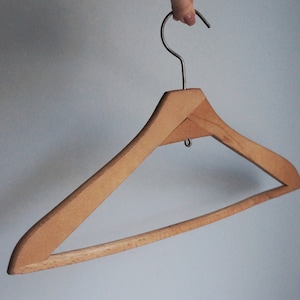 May include: A wooden clothes hanger with a metal hook. The hanger is made of light-colored wood and has a simple, classic design. The hook is silver-colored and attached to the top of the hanger.