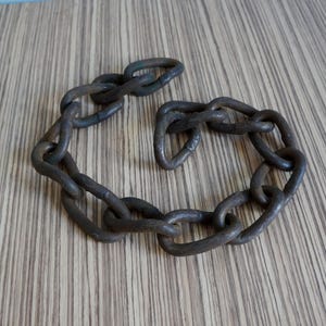 May include: A heavy-duty, dark metal chain with large, oval links. The chain is arranged in a circular shape, resting on a wooden surface with a striped pattern. The metal has a weathered appearance.