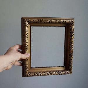 May include: A square, ornate gold-colored picture frame with detailed scrollwork. The frame is held against a plain gray background. The frame's interior is empty, ready for a photo or artwork.
