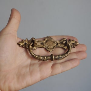 Op de afbeelding: Oud messing ladeknop met een sierlijk ontwerp. De knop heeft een centraal embleem en een gebogen handvat, met bloemaccenten aan elk uiteinde. De hardware heeft een warme goudkleur.