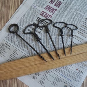 Op de afbeelding: Vier donkere metalen kurkentrekkers met T-vormige handvatten en spiraalvormige schachten. De gereedschappen zijn op een rij geplaatst, waarbij er één een cirkelvormig handvat heeft en drie T-vormige handvatten. De metalen oppervlakken zien er verouderd uit.