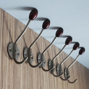 Könnte beinhalten: Sechs Vintage-Kleiderhaken an einer Holzwand befestigt. Jeder Haken hat einen silberfarbenen Metallstiel und einen dunkelbraunen, ovalen Holzknauf. Die Haken sind diagonal angeordnet und bieten ein klassisches Design.