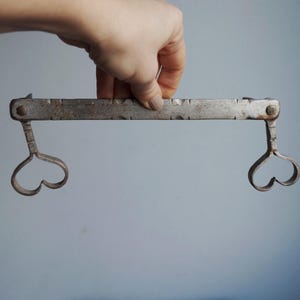 May include: An antique metal tool with a rectangular bar and heart-shaped ends. The bar has decorative notches along its length. The tool is held against a light blue background.
