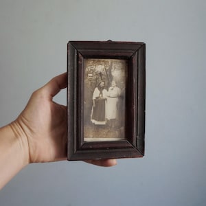 Könnte beinhalten: Ein kleiner, rechteckiger Bilderrahmen aus dunkelbraunem Holz mit einem alten Schwarz-Weiß-Foto von vier Personen, die im Freien stehen. Der Rahmen hat ein schlichtes Design mit abgeschrägten Kanten, sichtbarer Holzmaserung und einer abgenutzten Oberfläche.