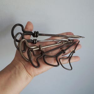以下が含まれることがあります： T字型のハンドルと尖ったシャフトを持つ5つの暗い金属製ツールのセット。各ツールは、ループ状または三角形のハンドルデザインと、まっすぐで先細りの金属棒が特徴です。金属の表面は、風化した暗い灰色の古びた質感を示しています。