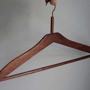 Puede incluir: Una percha de madera con un diseño curvo. La percha es de madera marrón oscuro y tiene un gancho de metal. La percha está diseñada para sujetar prendas de vestir y es un artículo doméstico común.